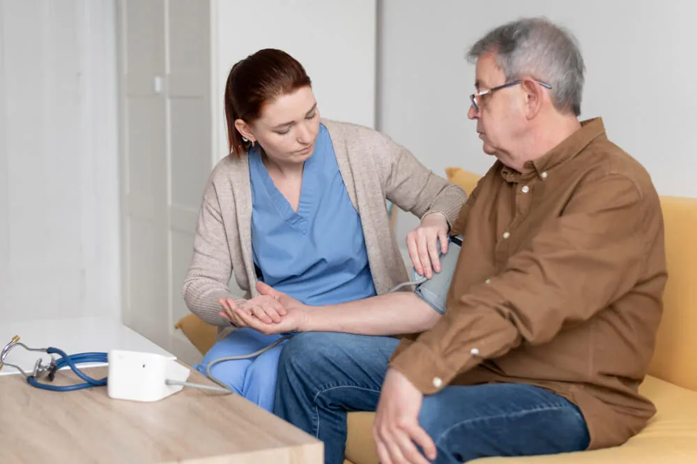A smiling clinician in green scrubs with a stethoscope rests a reassuring hand on the shoulder of an older female patient.