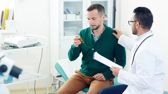 A doctor with a clipboard reassures a male patient who is examining a prescription bottle.