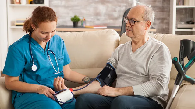 A nurse checks an older man’s blood pressure with a cuff while he sits on a sofa.